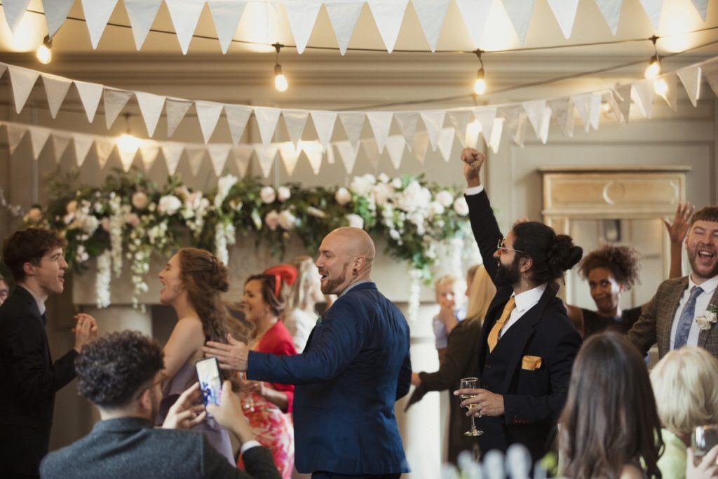 Wedding guests of mixed ages dressed smartly dancing at a wedding reception dancing with one another. One man is doing a fist pump and another is taking a video.