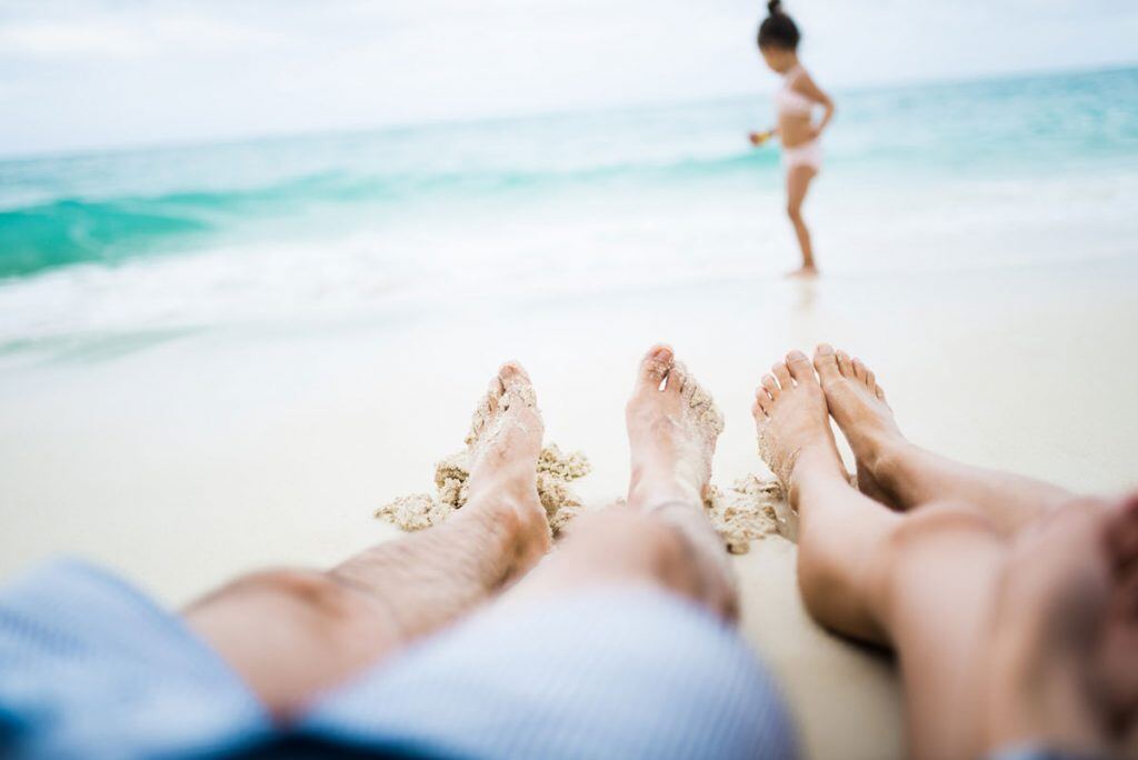 Parents taking a picture of their feet in the sand in the foreground and their daughter playing in the sand in the background | Motif