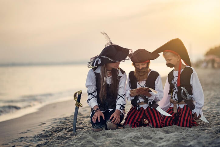 3 kids dressed as pirates read treasure map on the beach for Halloween photoshoot.