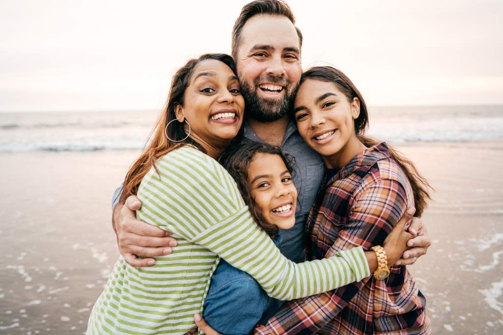 Mom and Dad with two girls on beach at sunset smile for a cute summer family picture.