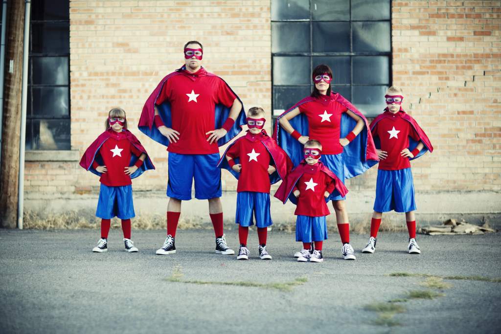 Parents with 4 small children pose by brick building in capes and masks for summer family picture.