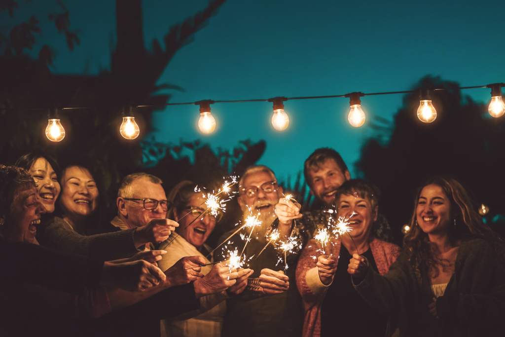 Happy family holds sparklers under string lights at night during backyard party.