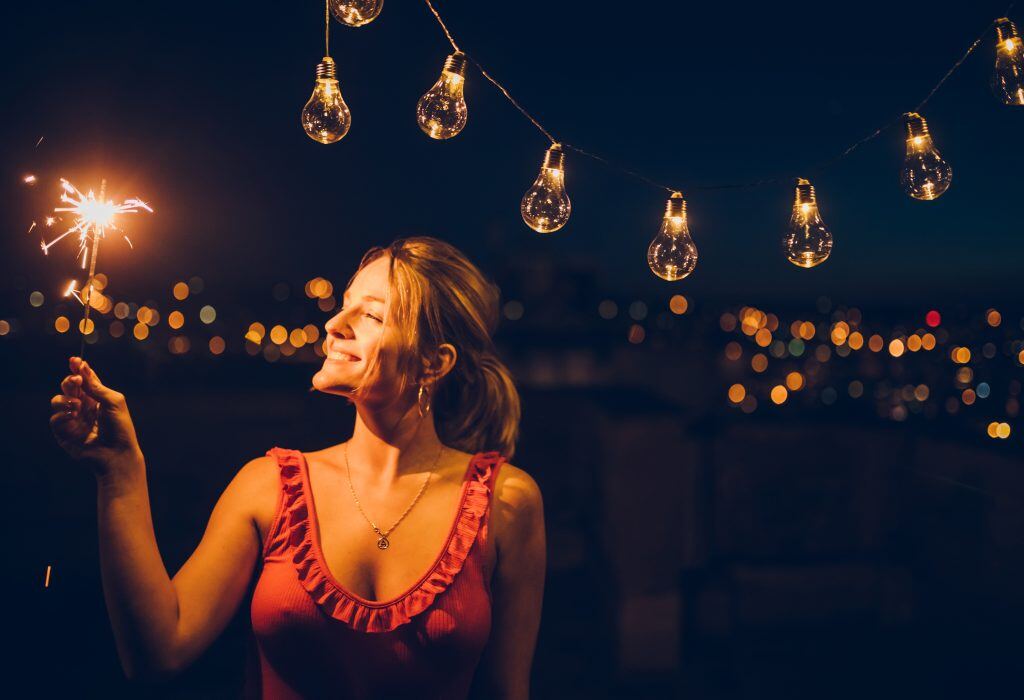 A sparkly summer photoshoot idea: woman with a sparkler standing under string lights.