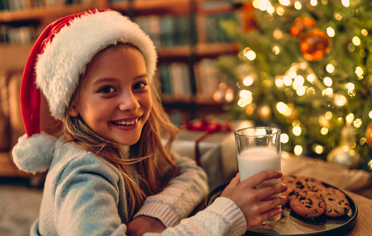 Young girl enjoys milk and cookies for Santa in front of the Christmas tree.  