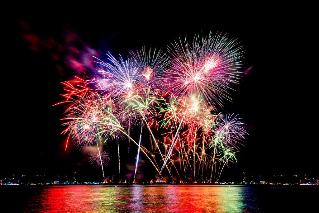 A colorful fireworks display over the beach reflects a rainbow in the water.