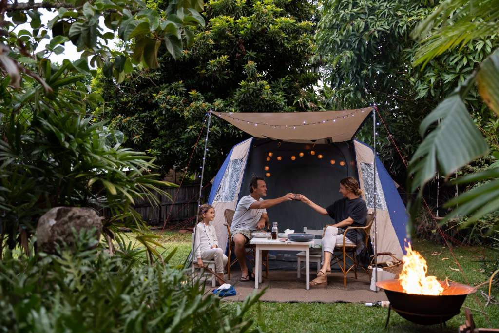 Family of three pitches tent in the backyard with fire pit and lights for summer family picture.