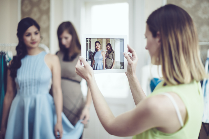 Young woman takes photography on iPad to capture young girl getting fitted for blue dress. 