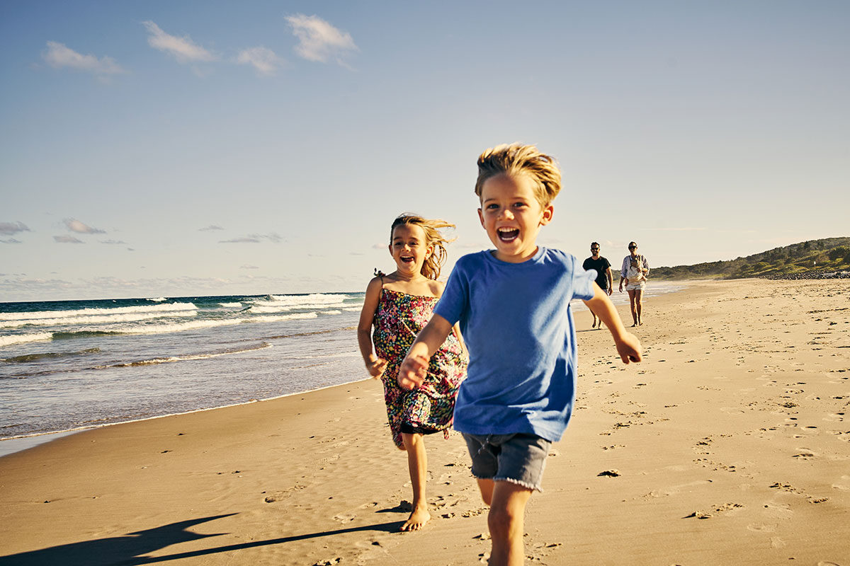 Two younger kids running on the beach with their parents walking behind | Motif<br />
