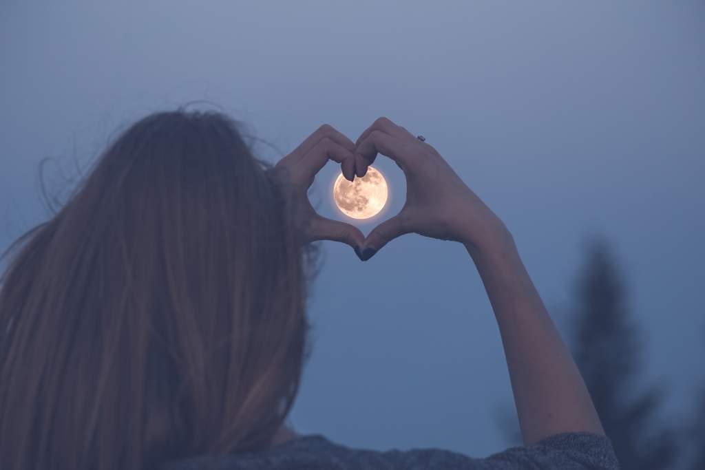 Woman uses hands to form a heart around the full moon in clear blue night sky.