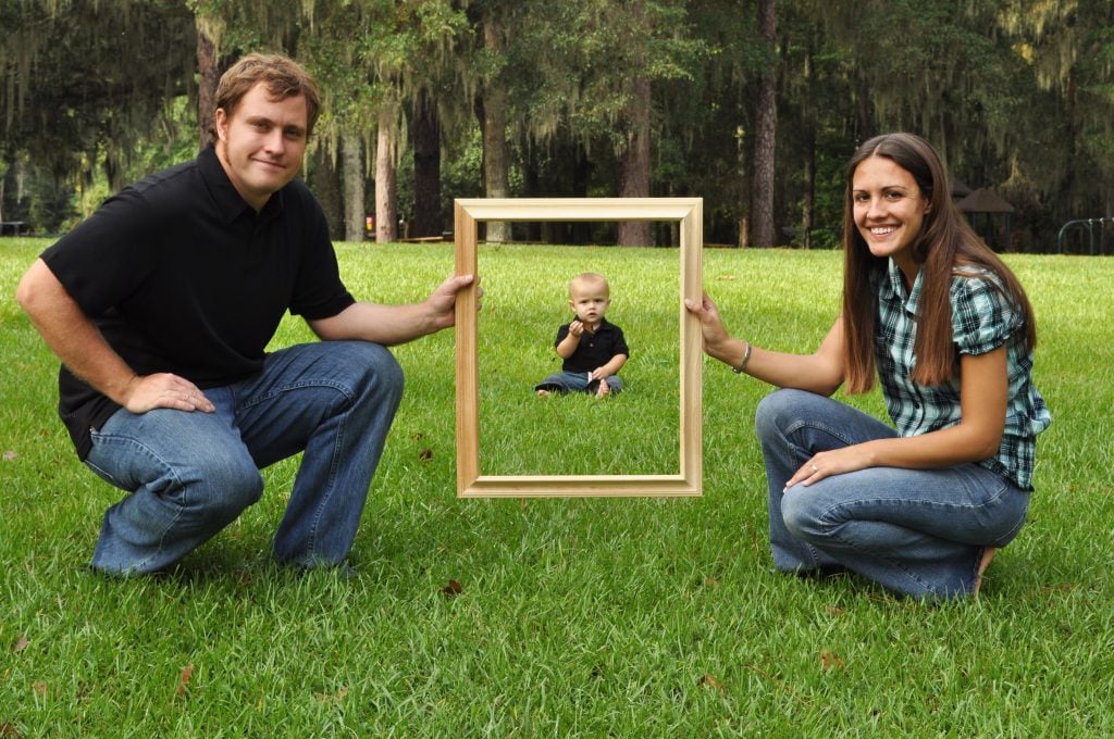 Mom and dad holding a picture frame with baby in background create forced perspective photography.